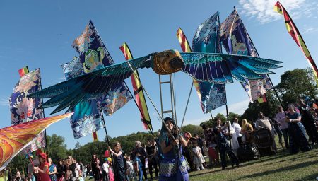 A performer operates a large, vibrant bird puppet with blue and teal wings during a sunny outdoor festival.