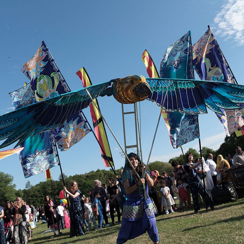A performer operates a large, vibrant bird puppet with blue and teal wings during a sunny outdoor festival.