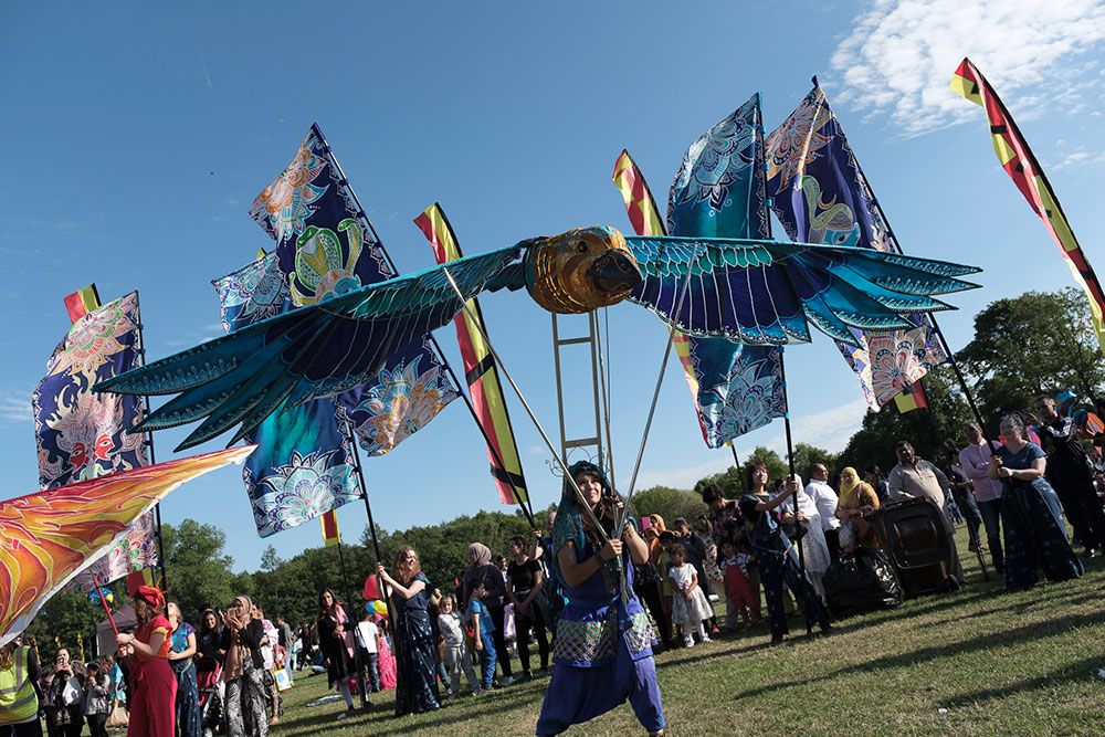 A performer operates a large, vibrant bird puppet with blue and teal wings during a sunny outdoor festival.