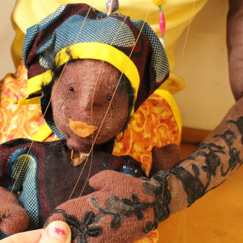 Close-up photo of a hand-crafted rod puppet with brown skin and textured, curly black hair wrapped in a colorful bandana. The puppet wears a patterned garment with floral and lace details on the sleeves.