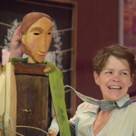 A smiling puppeteer performs with a large wooden puppet that has a long face and painted features. The puppet is draped in green fabric and held upright, while the performer looks off to the side with a joyful expression.