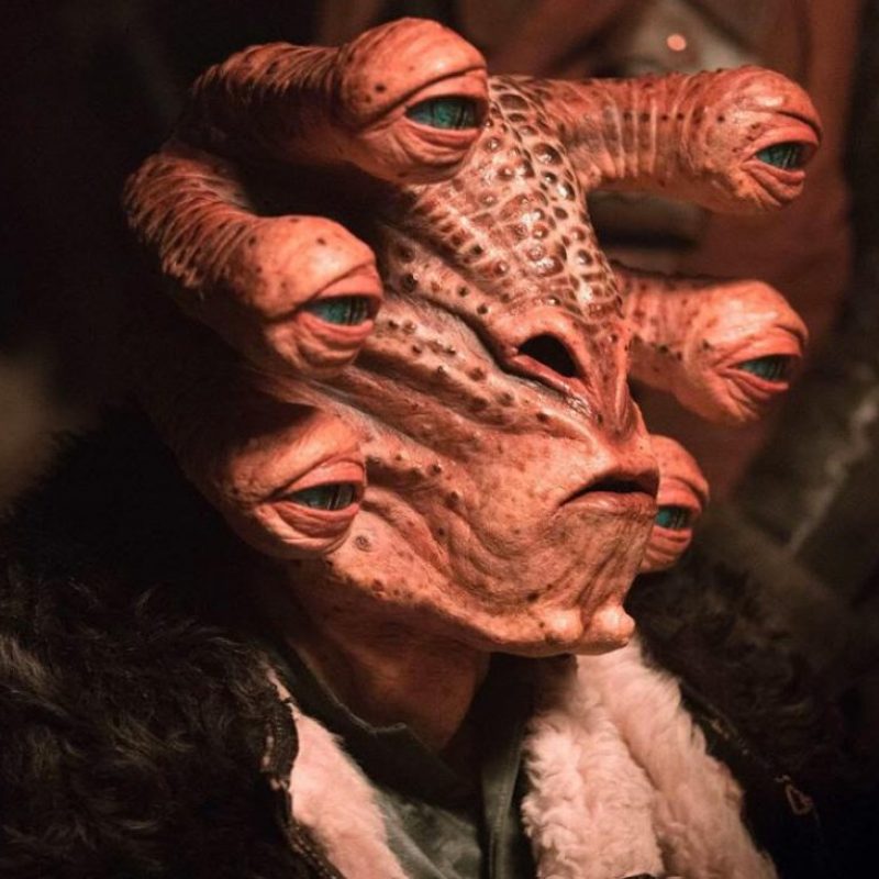 Close-up of a pinkish-orange alien with multiple eyes on thick stalks emerging from its head, wearing a dark fur-trimmed collar.