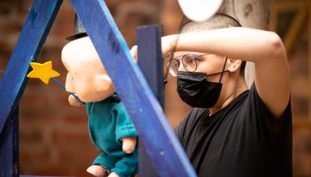 GPT A person with glasses and a face mask attentively adjusts a puppet on a blue wooden stand