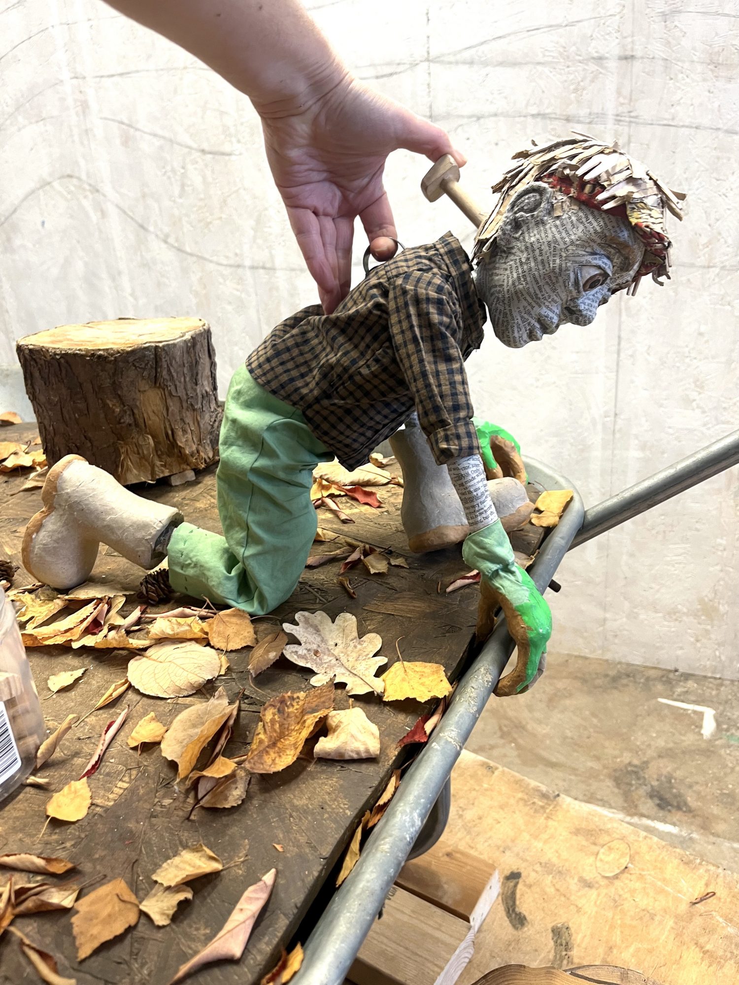 A hand manipulates a papier-mâché puppet to lean over the edge of a wheelbarrow filled with autumn leaves, next to a small wooden stump.