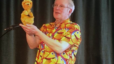 'A puppeteer on stage holds a small yellow lion puppet while performing in front of a microphone.