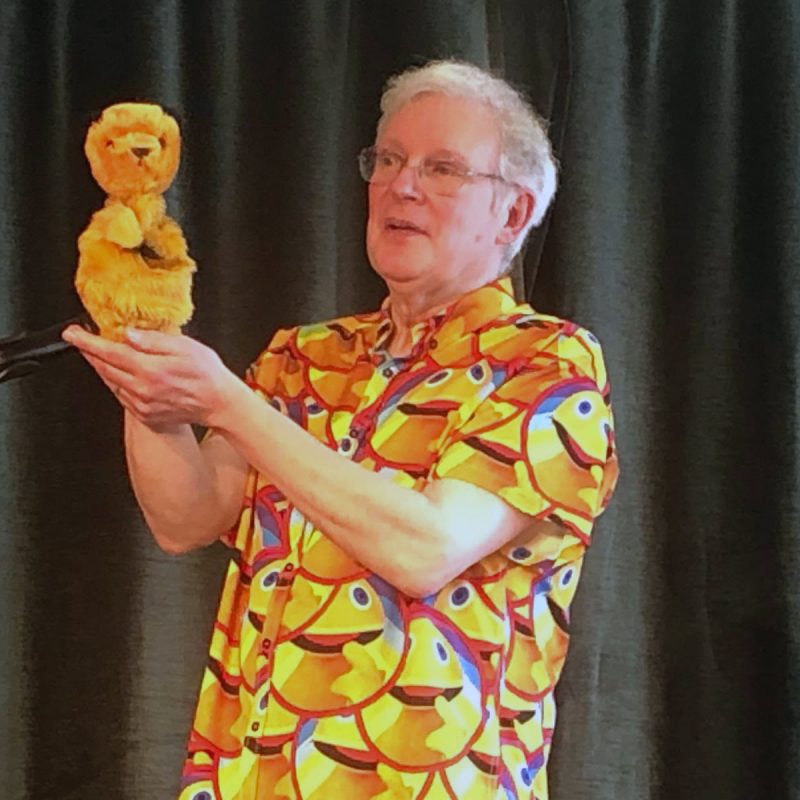 'A puppeteer on stage holds a small yellow lion puppet while performing in front of a microphone.