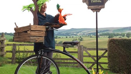 A puppeteer stands in a field alongside a penny farthing, holding a small orange puppet