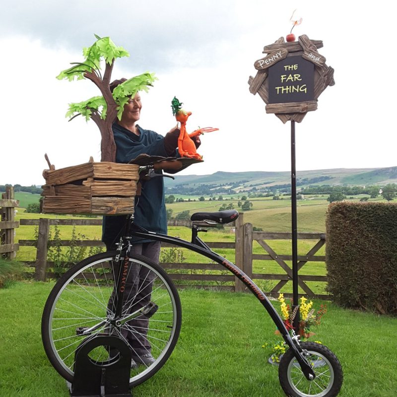 A puppeteer stands in a field alongside a penny farthing, holding a small orange puppet