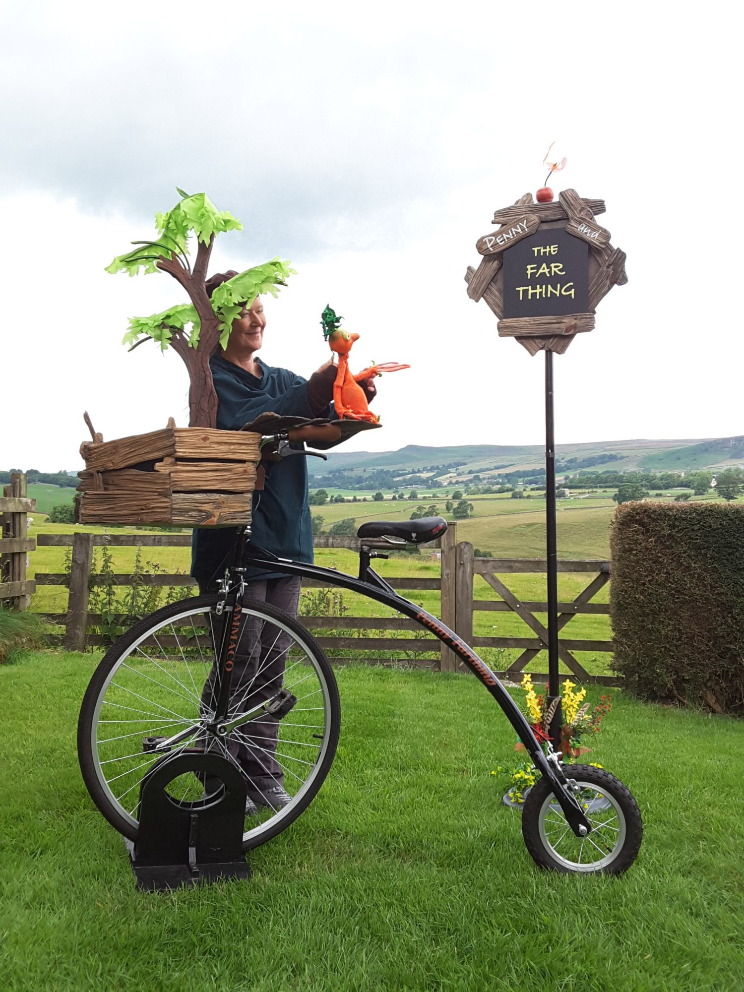 A puppeteer stands in a field alongside a penny farthing, holding a small orange puppet