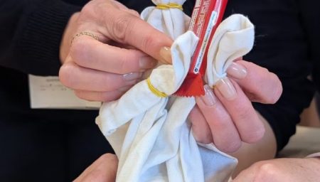 A person’s hands assembling a simple puppet from white cloth tied with yellow rubber bands, with a red candy wrapper tucked into the fabric.