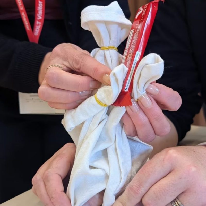 A person’s hands assembling a simple puppet from white cloth tied with yellow rubber bands, with a red candy wrapper tucked into the fabric.