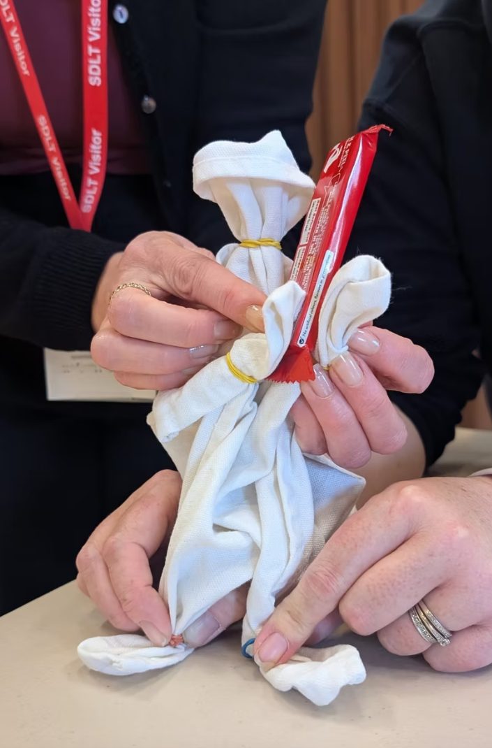 A person’s hands assembling a simple puppet from white cloth tied with yellow rubber bands, with a red candy wrapper tucked into the fabric.