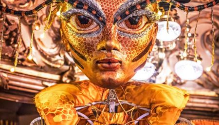 A close-up of a large, golden puppet head with human and bee features, wearing a decorated top hat.
