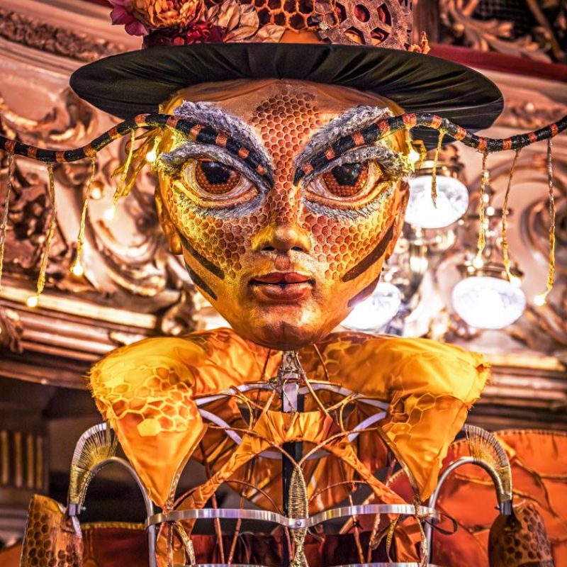 A close-up of a large, golden puppet head with human and bee features, wearing a decorated top hat.