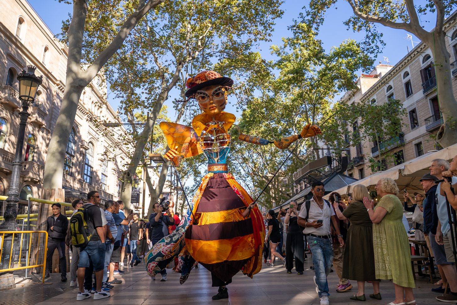 A tall, illuminated "Queen Bee" puppet with a golden face and a decorated top hat walks through crowds at an outdoor festival.