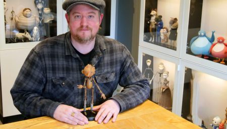 A man in a flat cap and plaid shirt sits at a wooden table with a wooden Pinocchio puppet, surrounded by glass display cases filled with stop-motion animation characters.