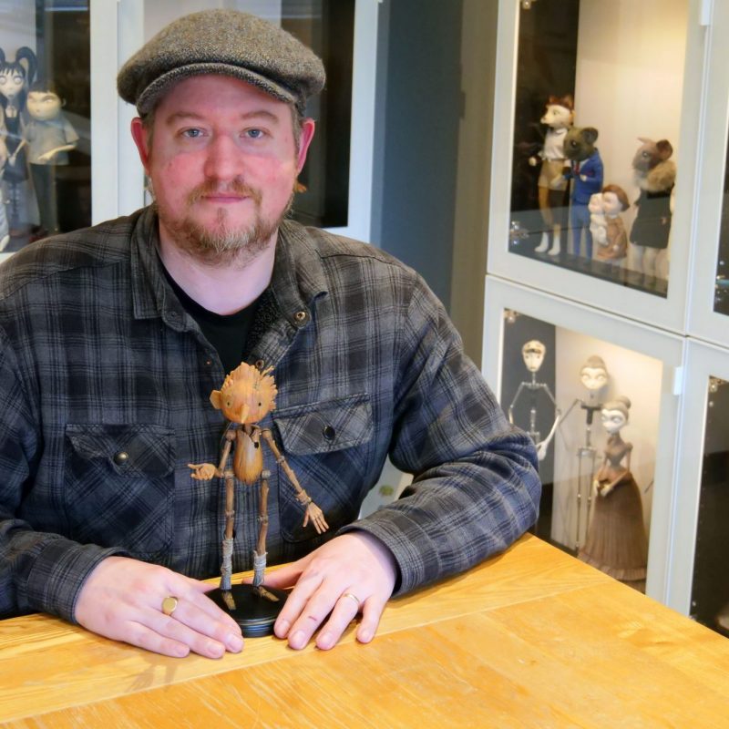 A man in a flat cap and plaid shirt sits at a wooden table with a wooden Pinocchio puppet, surrounded by glass display cases filled with stop-motion animation characters.