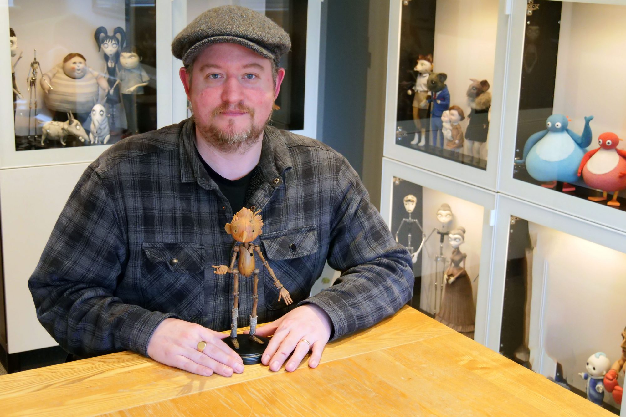 A man in a flat cap and plaid shirt sits at a wooden table with a wooden Pinocchio puppet, surrounded by glass display cases filled with stop-motion animation characters.