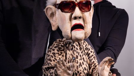 An elderly woman puppet with white fuzzy hair, oversized sunglasses, and a leopard-print cardigan is being operated by two puppeteers in black clothing