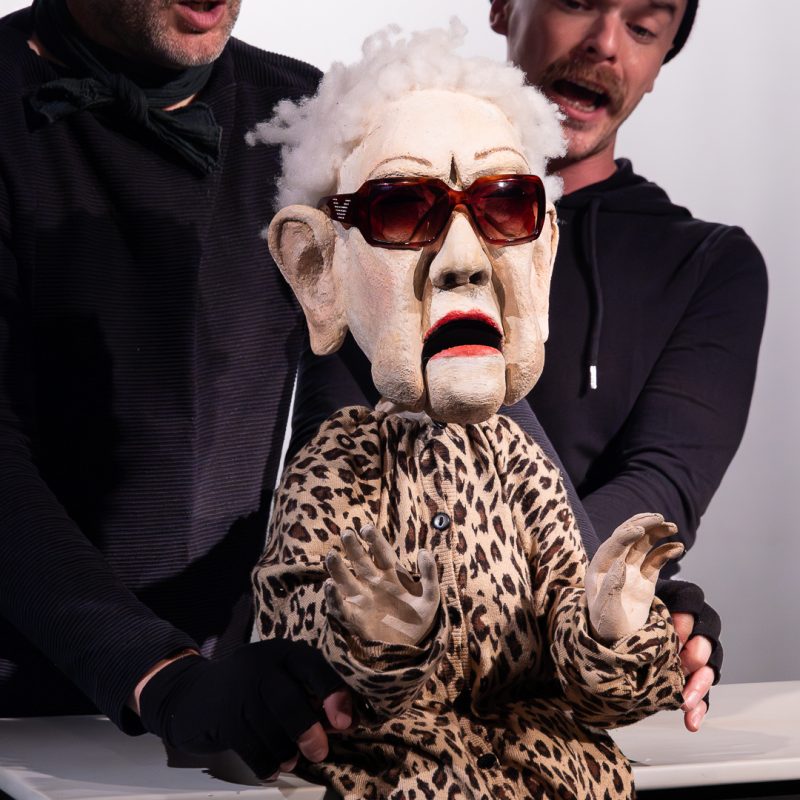 An elderly woman puppet with white fuzzy hair, oversized sunglasses, and a leopard-print cardigan is being operated by two puppeteers in black clothing