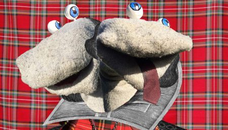 A two-headed grey sock puppet wearing an Elizabethan doublet and ruff collar, positioned against a red plaid background.