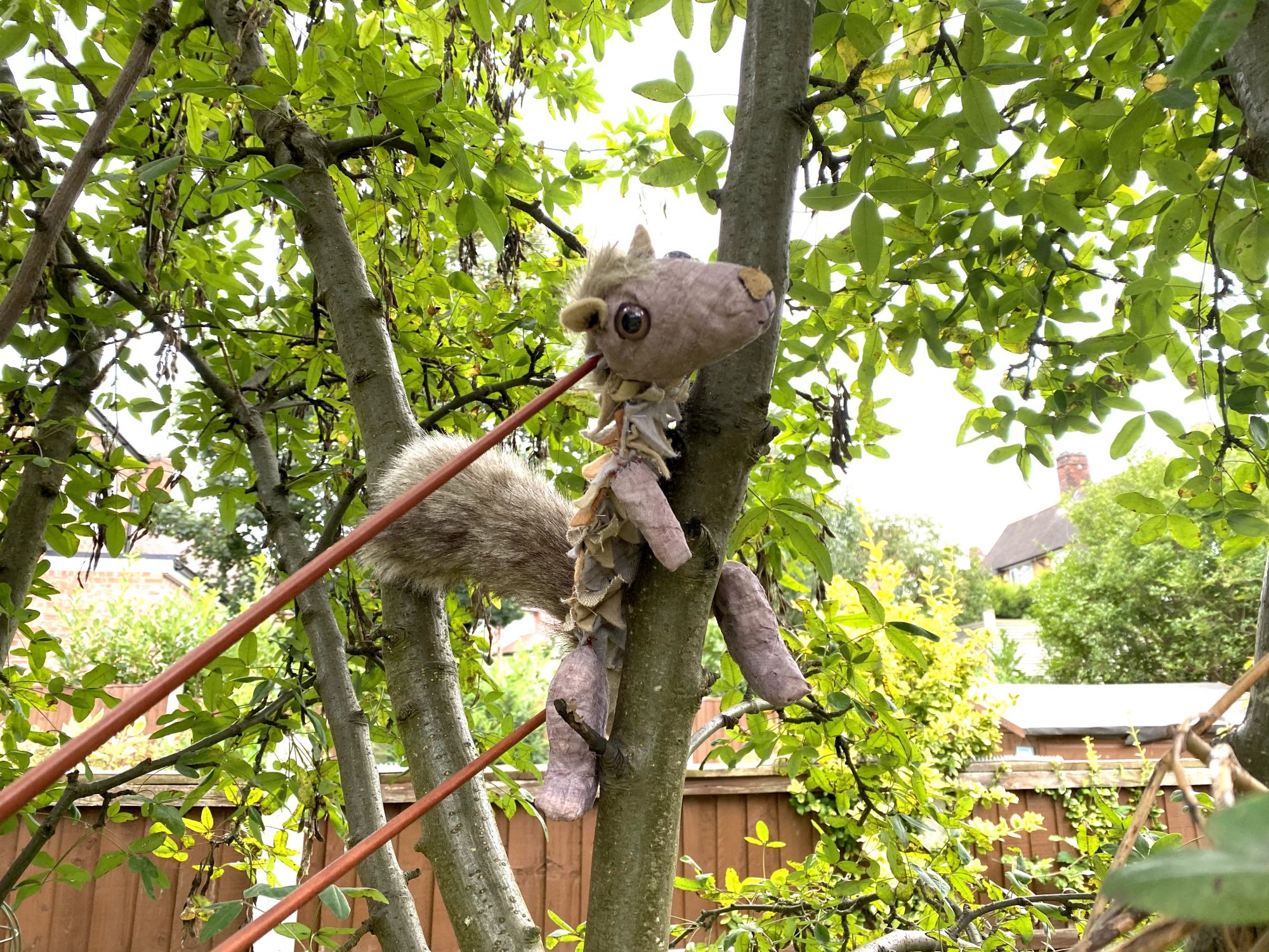 A grey squirrel puppet with a bushy tail and large glass eyes clinging to the trunk of a leafy green tree. Two thin orange control rods are attached to the puppet, operated from below. The background shows a sunny garden with a wooden fence and distant houses.