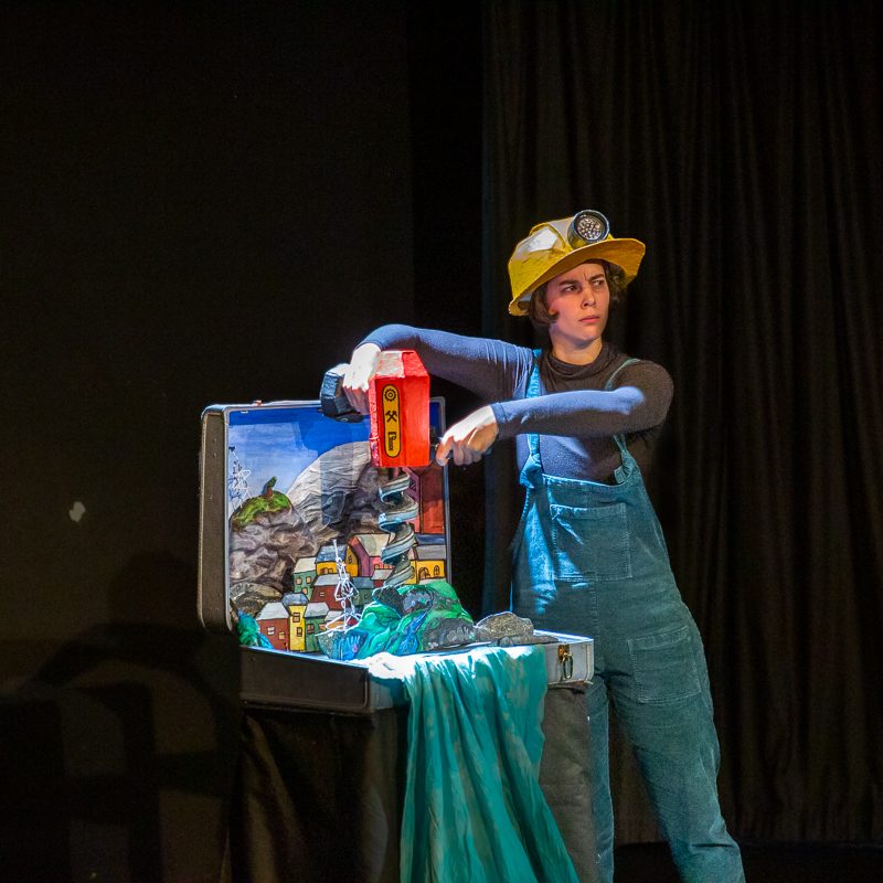 An actor in a yellow hardhat and blue overalls uses a prop drill on a miniature mountain landscape built inside an open suitcase on a stage.