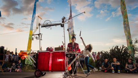A street performer in a red checkered jacket stands behind a complex, Rube Goldberg-style contraption on wheels while a young girl reaches up to interact with it.