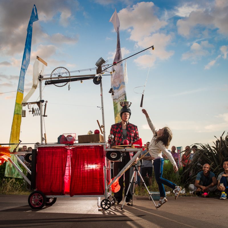 A street performer in a red checkered jacket stands behind a complex, Rube Goldberg-style contraption on wheels while a young girl reaches up to interact with it.