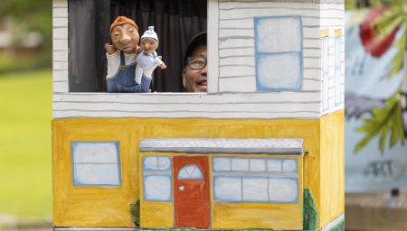 A puppeteer peeks out from a mobile puppet stage shaped like a two-story house, holding two small hand puppets in the top window.