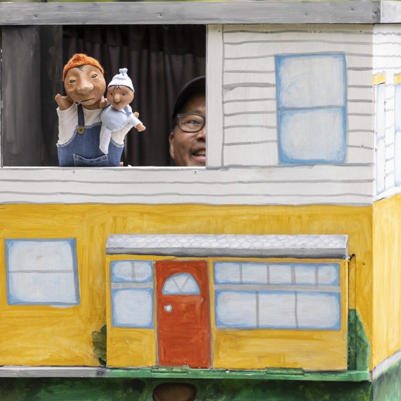 A puppeteer peeks out from a mobile puppet stage shaped like a two-story house, holding two small hand puppets in the top window.