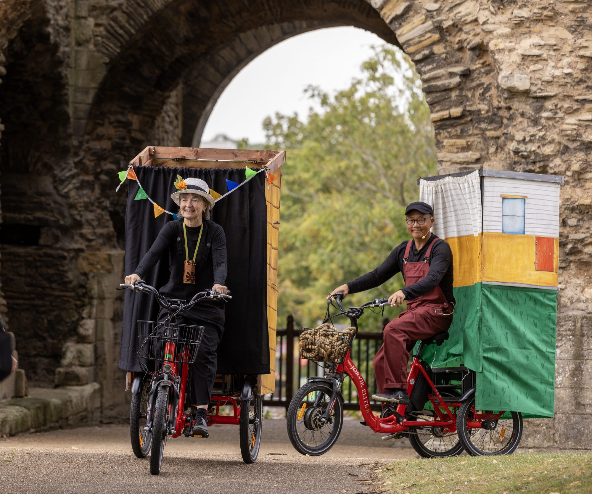 Two performers ride red tricycles carrying mobile puppet stages through a large stone archway.