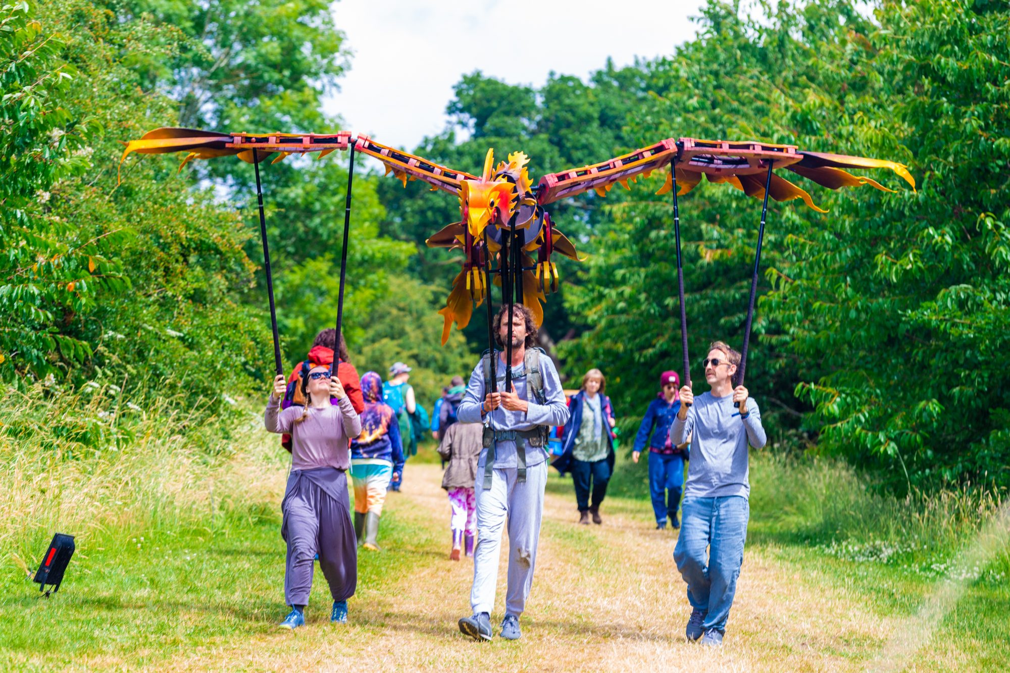 Three performers walking along a grassy path outdoors, operating a large, mechanical phoenix puppet with wide orange wings