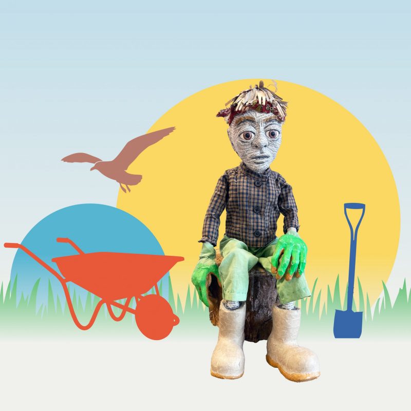 A stylised promotional image featuring a handmade puppet seated on a tree stump. The puppet has large, expressive eyes, a face made from printed text, short straw-like hair, green gloves, and wears a checked shirt, green trousers and white boots. Behind it are simple graphic shapes: a large yellow circle, a smaller blue circle, grass silhouettes, and icons including a red wheelbarrow, a blue spade, and a bird in flight.