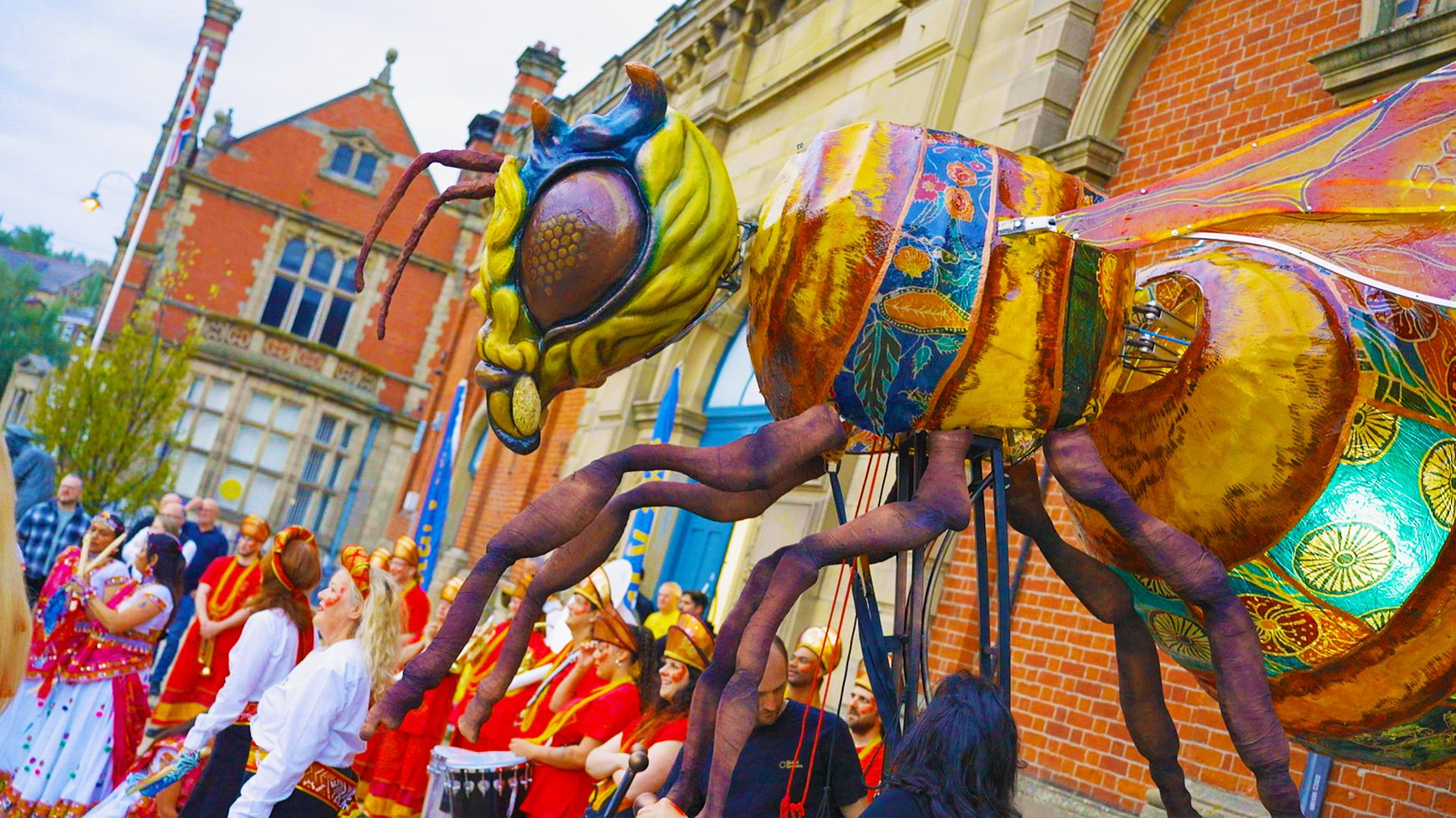 A massive, brightly patterned mechanical bee puppet is paraded past a brick building during a street festival.