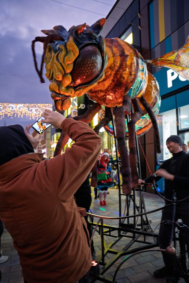 A man takes a photo of a giant, illuminated worker bee puppet during an evening street parade.