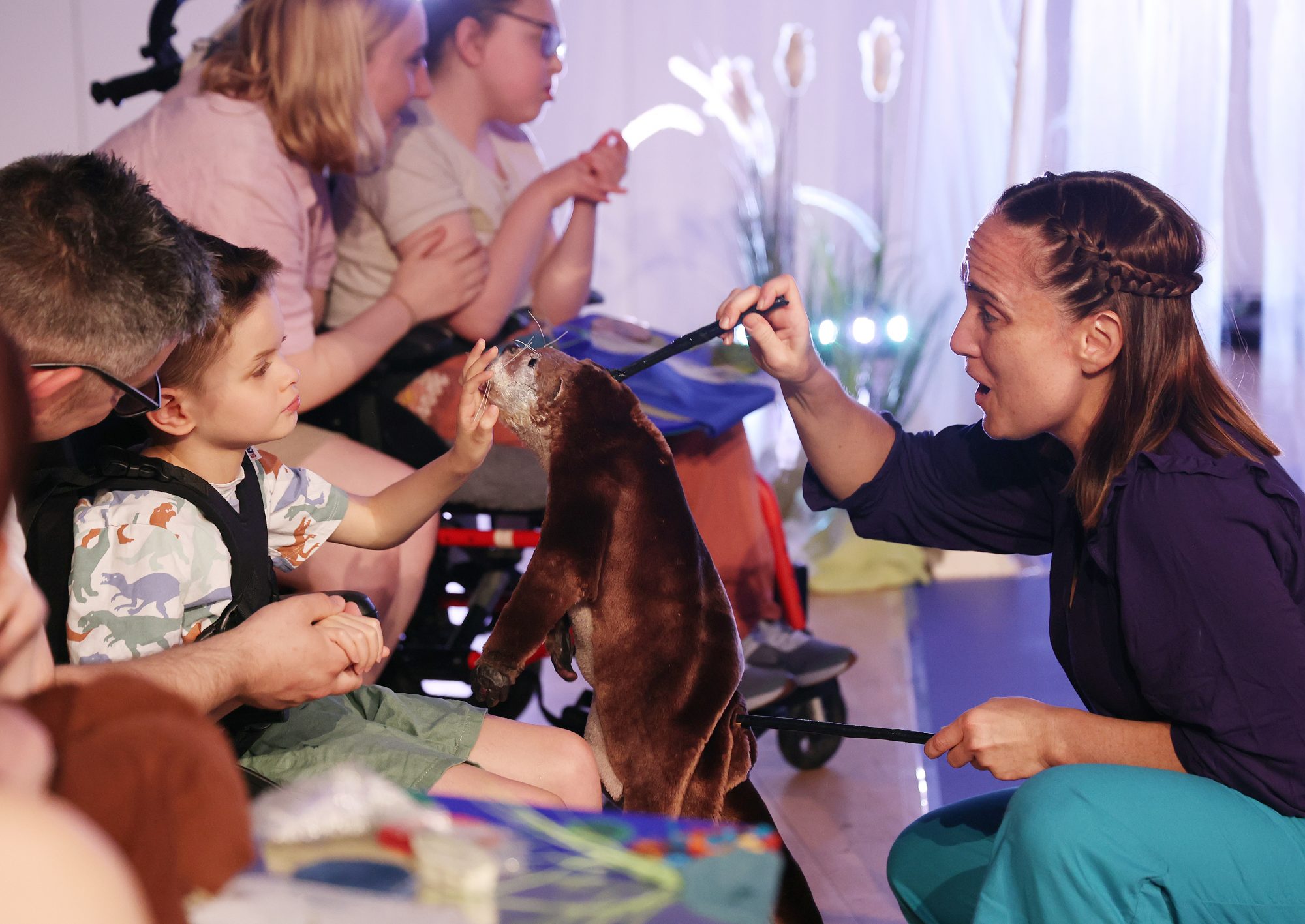 A performer uses rods to move a realistic brown otter puppet toward a young boy, who reaches out to touch its nose.