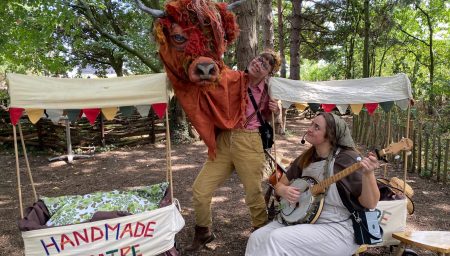 Two performers in a wooded area; one operates a large, shaggy highland cow puppet while the other sits and plays a banjo next to a 