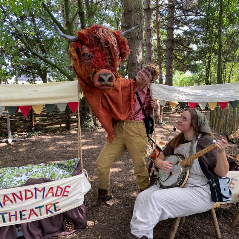 Two performers in a wooded area; one operates a large, shaggy highland cow puppet while the other sits and plays a banjo next to a 