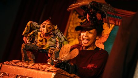 A puppeteer with an animated expression performs with a wide-eyed, spindly-limbed puppet over an open wooden trunk on a dimly lit stage.