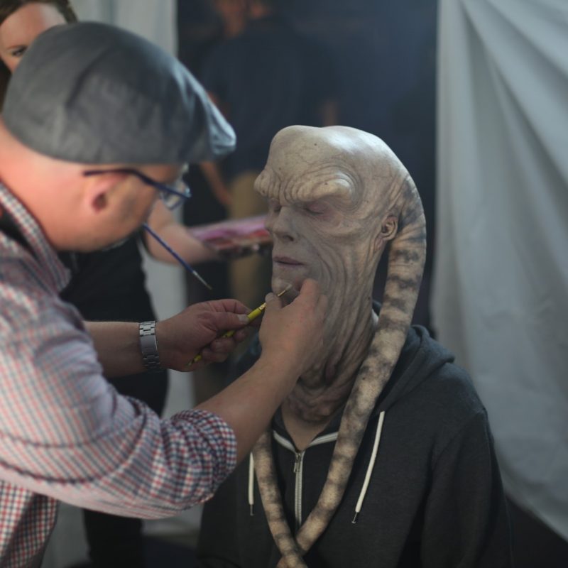 A makeup artist is applying finishing touches to a person in an elaborate alien prosthetic mask for a film production